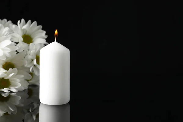 Burning candle and white chrysanthemum flowers on black mirror surface in darkness, space for text. Funeral symbols