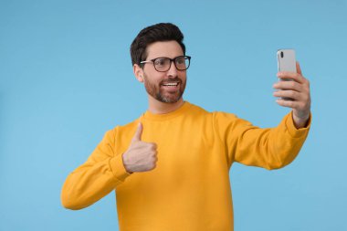 Gülümseyen adam akıllı telefonuyla selfie çekiyor ve açık mavi arka planda baş parmak gösteriyor.