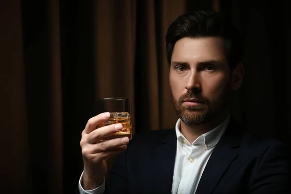 Man in suit holding glass of whiskey with ice cubes on brown background