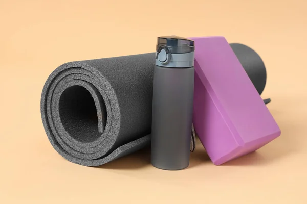 Grey exercise mat, yoga block and bottle of water on beige background