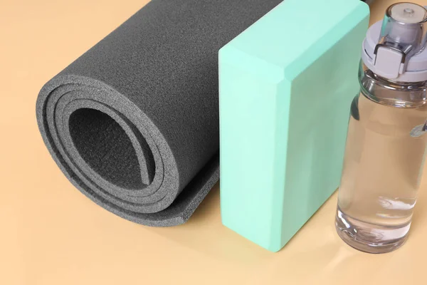 Grey exercise mat, yoga block and bottle of water on beige background, closeup