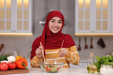 Müslüman kadın, mutfaktaki beyaz masada sebzelerle lezzetli salata yapıyor.