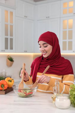 Müslüman kadın, mutfaktaki beyaz masada sebzelerle lezzetli salata yapıyor.