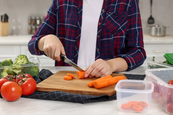 Woman cutting carrot near containers with fresh products on white marble table in kitchen, closeup. Food storage