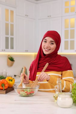 Müslüman kadın, mutfaktaki beyaz masada sebzelerle lezzetli salata yapıyor.