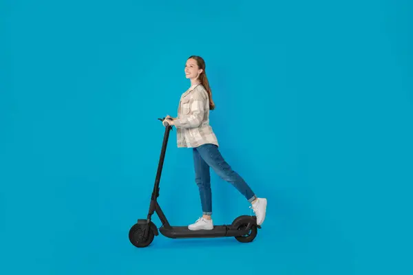 ライトブルーの背景にモダンな電気キックスクーターに乗っている幸せな女性 — ストック写真