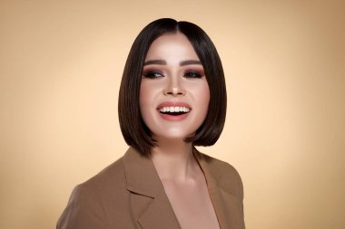 Bej arka planda kahverengi saçlı ve gülümseyen zarif genç bir kadının portresi.