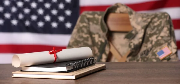 Military education. Notebooks and diploma on wooden table, chair with soldier\'s jacket against flag of United States indoors