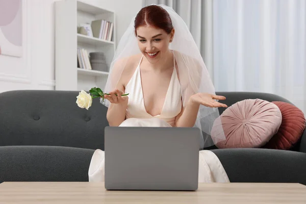 Happy bride with rose having online video chat via laptop in living room