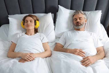 Evde, horlayan kocasının yanında uzanmış kulaklıklı, gülümseyen bir kadın.