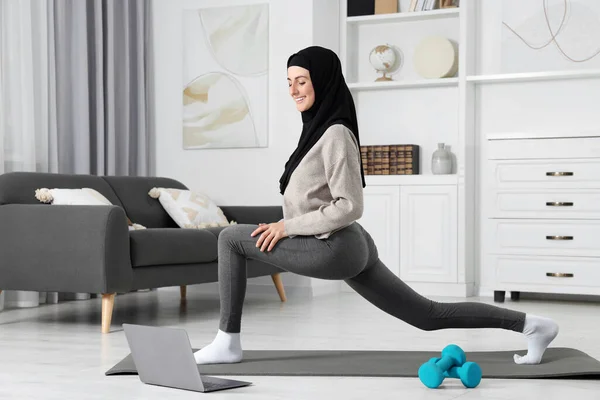 Muslim woman in hijab stretching near laptop on fitness mat at home