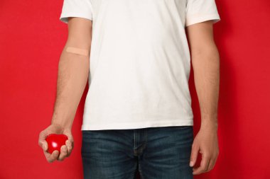 Kan bağışı konsepti. Kolunda yapıştırıcı olan adam kırmızı kalbi arka planda tutuyor, yakın plan.