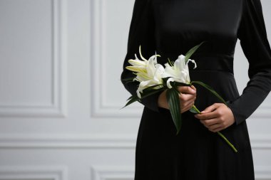 Woman with lilies near white wall, closeup and space for text. Funeral ceremony clipart