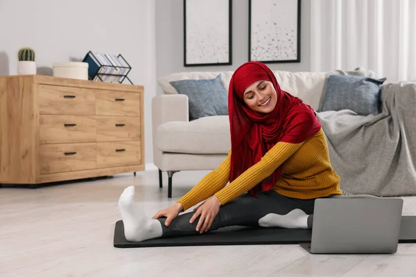 Muslim woman in hijab stretching near laptop on fitness mat at home. Space for text