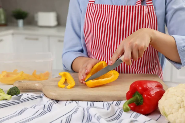 Woman cutting bell pepper near food storage container at table in kitchen, closeup