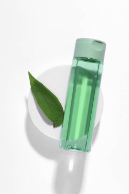 Beyaz arka planda bir şişe kozmetik ürünü, yeşil yaprak ve podyum.