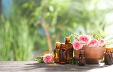 Bottles with essential oils, roses and rosemary on wooden table against blurred green background. Space for text clipart