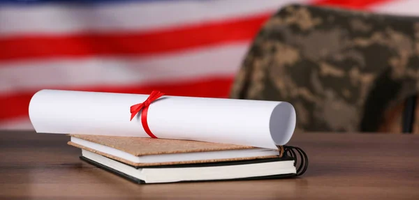 Military education. Diploma and notebooks on wooden table against flag of USA. Chair with soldier\'s jacket indoors