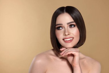 Kahverengi saçlı, bej arka planda gülümseyen güzel bir genç kadının portresi.