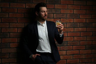 Resmi kıyafetli bir adam kırmızı tuğla duvarın yanında viski bardağı tutuyor.