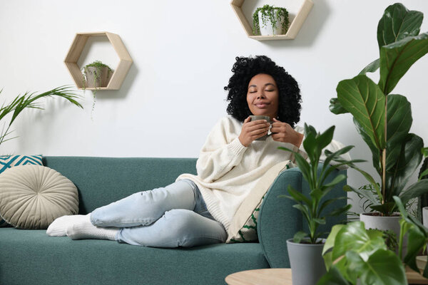 Relaxing atmosphere. Woman with cup of hot drink on sofa near beautiful houseplants in room