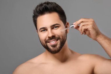 Gri arka planda yüzüne kozmetik serumu süren yakışıklı bir adam.