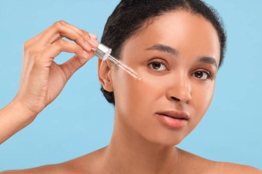 Açık mavi arka planda yüzüne serum süren güzel bir kadın. Yakın plan.