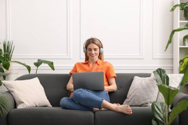 Kulaklıklı bir kadın evdeki güzel saksıların yanındaki kanepede dizüstü bilgisayar kullanıyor.