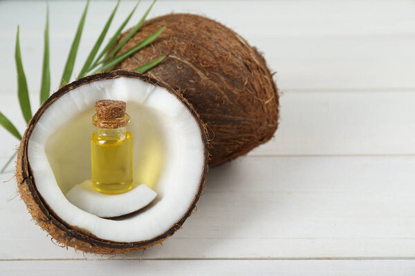 Bottle of organic coconut cooking oil, leaf and fresh fruits on white wooden table, space for text