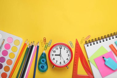 Sarı zemin üzerinde farklı okul kırtasiyesi ve alarm saati olan düz kompozisyon, mesaj için boşluk. Okula geri dön.