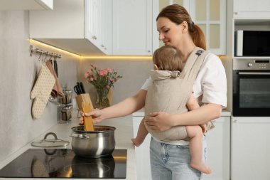 Anne, mutfakta makarna pişirirken çocuğunu askıda tutuyor.