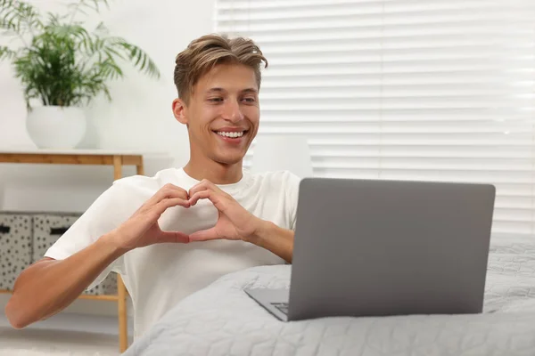 Happy young man having video chat via laptop and making heart with hands on bed indoors. Long-distance relationship