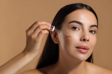 Güzel genç bir kadın bej arka planda yüzüne serum sürüyor.