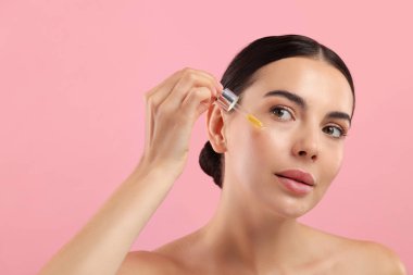 Güzel genç bir kadın pembe arka planda yüzüne serum sürüyor.