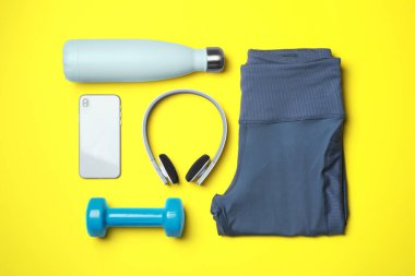 Şık spor taytları, dambıllar, termo şişeler, kulaklıklar ve sarı arka planda akıllı telefonlar.