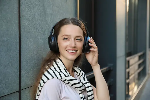 Smiling woman in headphones listening to music near building outdoors