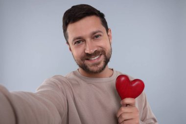 Kırmızı kalbi tutan ve açık gri arkaplanda selfie çeken mutlu bir adam.