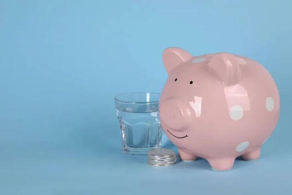Water scarcity concept. Piggy bank, coins and glass of drink on light blue background, space for text