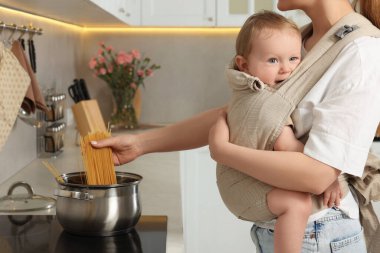 Anne, mutfakta makarna pişirirken çocuğunu askıda tutuyor.