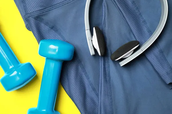 Stylish sports leggings, headphones and dumbbells on yellow background, flat lay