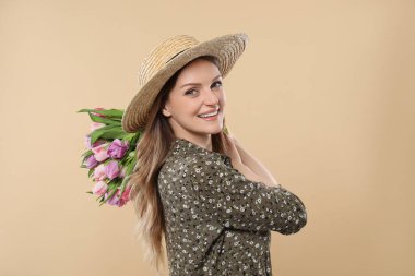 Hasır şapkalı mutlu genç kadın elinde bej arka planda güzel laleler tutuyor.