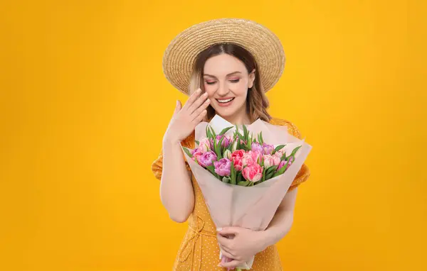 黄色の背景に美しいチューリップの花束を保持するわら帽子で幸せな若い女性 — ストック写真