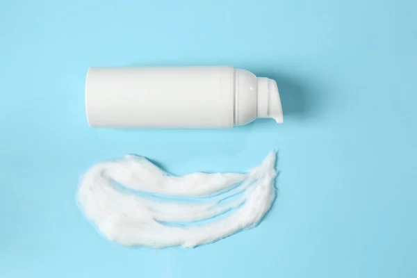 Bottle of face cleanser and white foam on light blue background, top view. Skin care cosmetic
