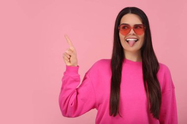 Kalp şeklinde gözlüklü, dilini gösteren ve pembe arka planı işaret eden mutlu genç bir kadın. Metin için boşluk