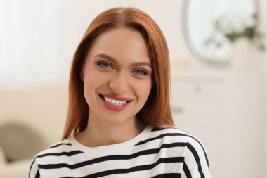 Portrait of beautiful young woman with red hair at home. Attractive lady smiling and looking into camera clipart