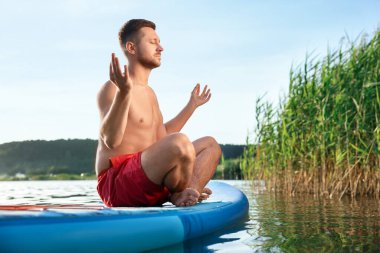 Adam gün batımında nehirde açık mavi SUP tahtası üzerinde meditasyon yapıyor.