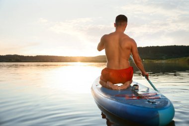 Günbatımında nehrin arka tarafında, SUP tahtasının üzerinde kürek çeken bir adam.