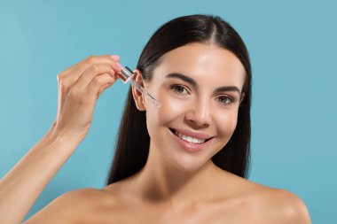 Güzel genç bir kadın açık mavi arka planda yüzüne serum sürüyor.