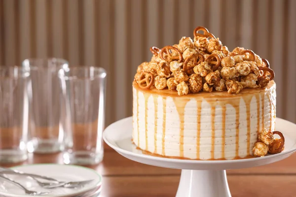 Caramel drip cake decorated with popcorn and pretzels near tableware on table