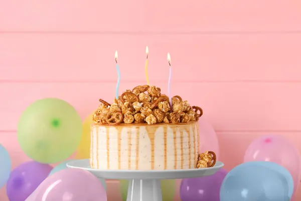 Caramel drip cake decorated with popcorn and pretzels near balloons against pink wooden background